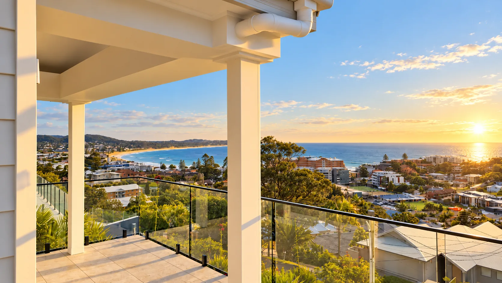 Sunrise over the ocean from a covered balcony at The Terrigal Residence, with white columns and a frameless glass balustrade, looking over treetops and rooftops to a curving beach, the ocean and the sun low on the horizon