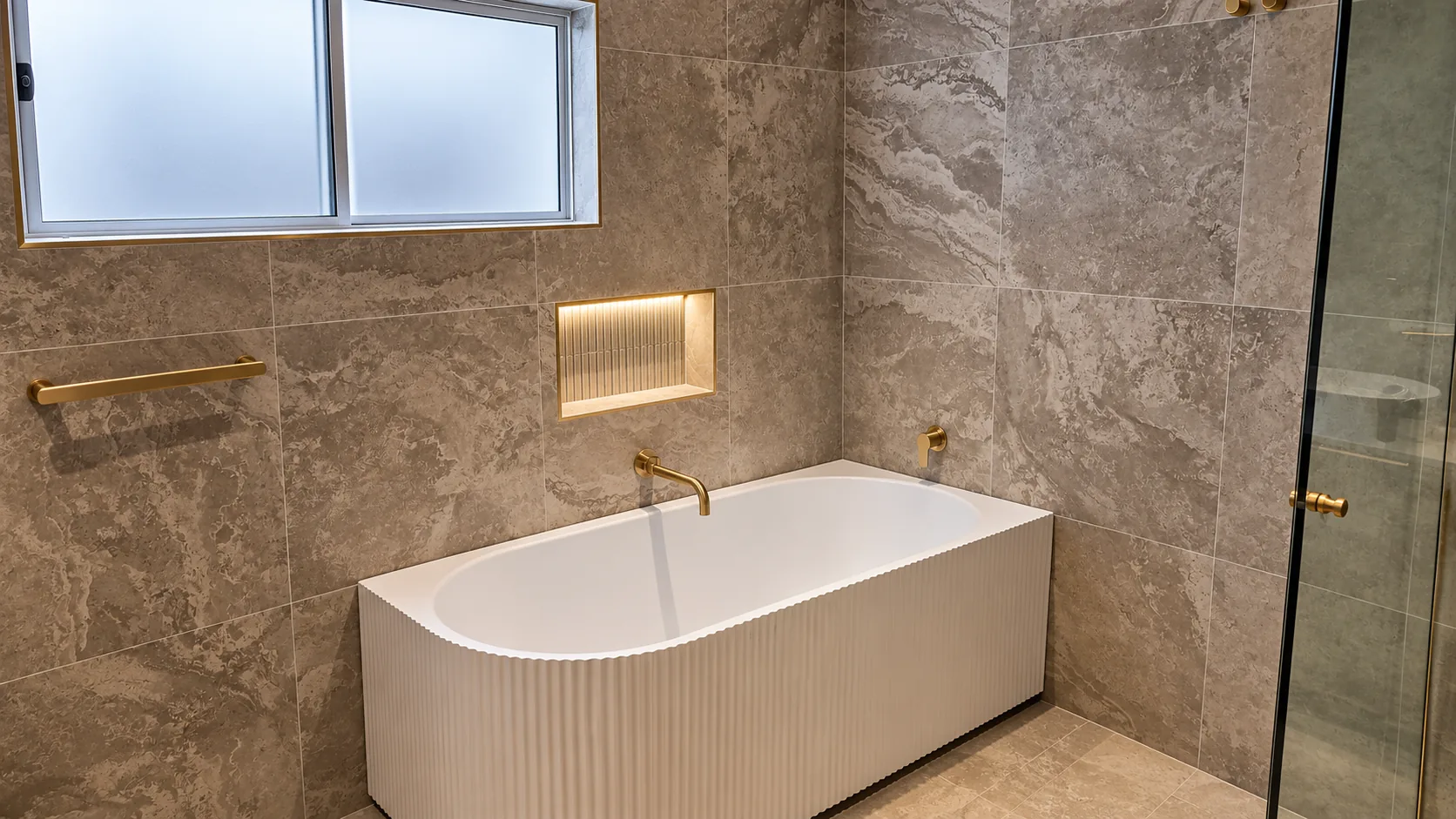 Fluted white bathtub with a wall-mounted brass-toned spout, a lit recessed niche with fine vertical tiles, a brass-toned towel rail and large stone-look tiles in warm taupe