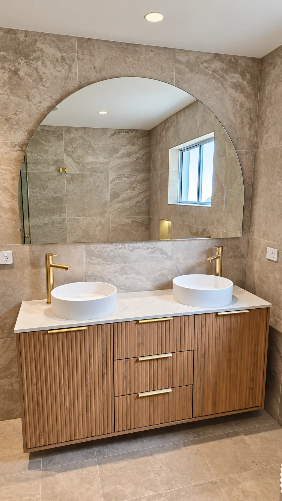 Bathroom vanity with ribbed timber doors and drawers, brass-toned handles, a white stone top carrying twin round basins with tall brass-toned mixers, and a large arched mirror set against stone-look wall tiles