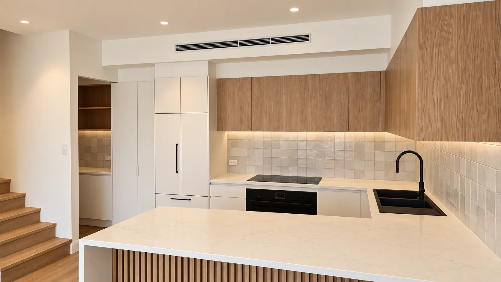 Contemporary kitchen in the Terrigal residence, with timber-finish overhead cabinetry above a softly lit tiled splashback, white tall cabinetry with an integrated refrigerator, black cooktop, oven and sink, and a stone-topped peninsula with a slatted timber base