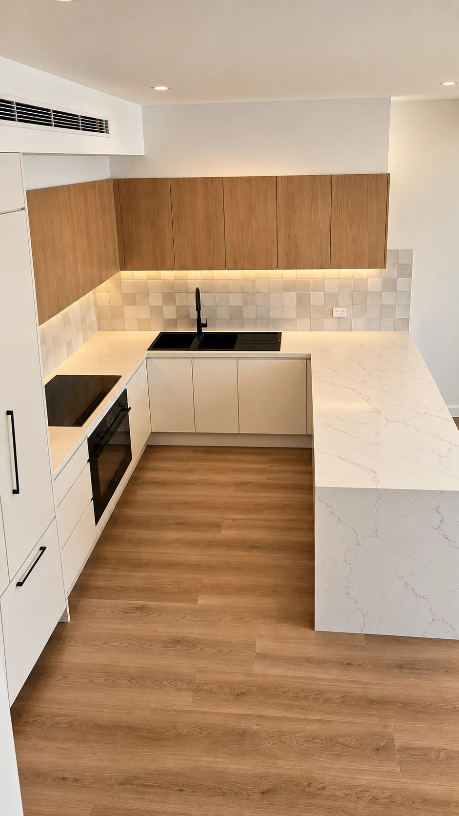 U-shaped kitchen seen from above, with a veined stone benchtop that falls to the floor in a waterfall edge, handleless white base cabinets, a black sink and tapware, and timber overhead cabinetry with under-cabinet lighting