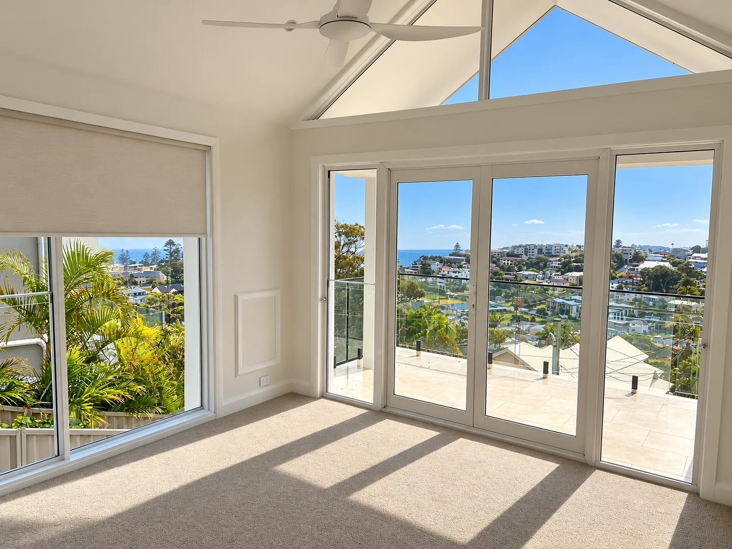 Terrigal master bedroom with balcony and ocean views: a raked ceiling, triangular highlight windows and a ceiling fan, sunlit carpet, a large window with a roller blind framing palms, and glazed doors opening to a tiled balcony with a glass balustrade and views over rooftops to the ocean