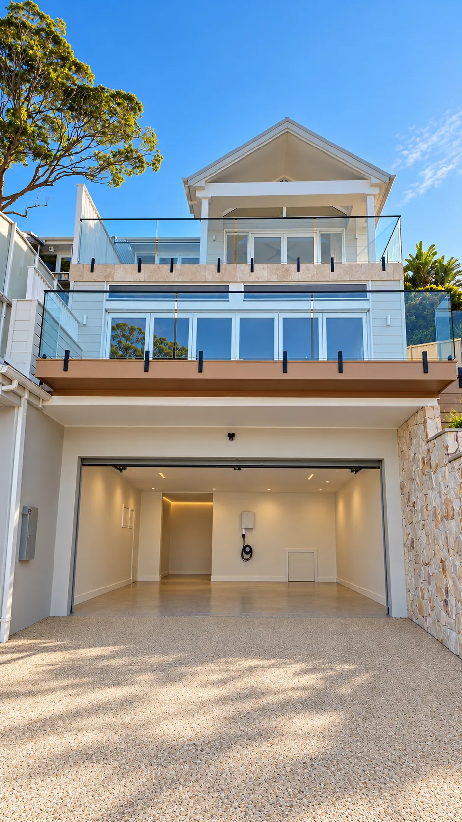 Front of the Terrigal residence under a clear blue sky: an open double garage with a lit interior at street level, three levels of balconies with glass balustrades above, rising to a gabled roof, beside a sandstone wall