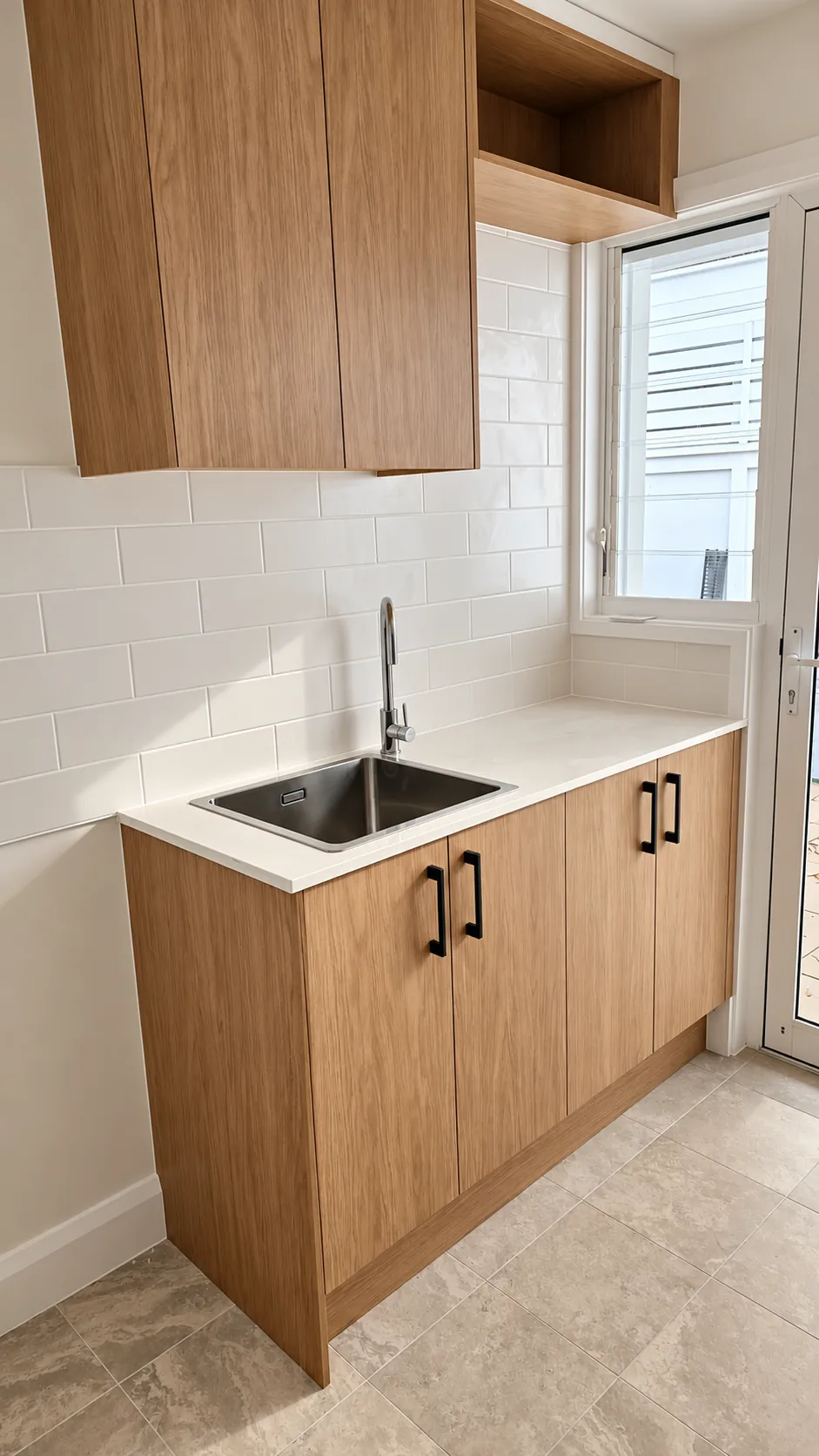 Laundry with timber cabinetry and black bar handles, a stainless steel sink in a white benchtop, white subway-tile splashback, an open timber shelf above, and a window and glazed door to the outside