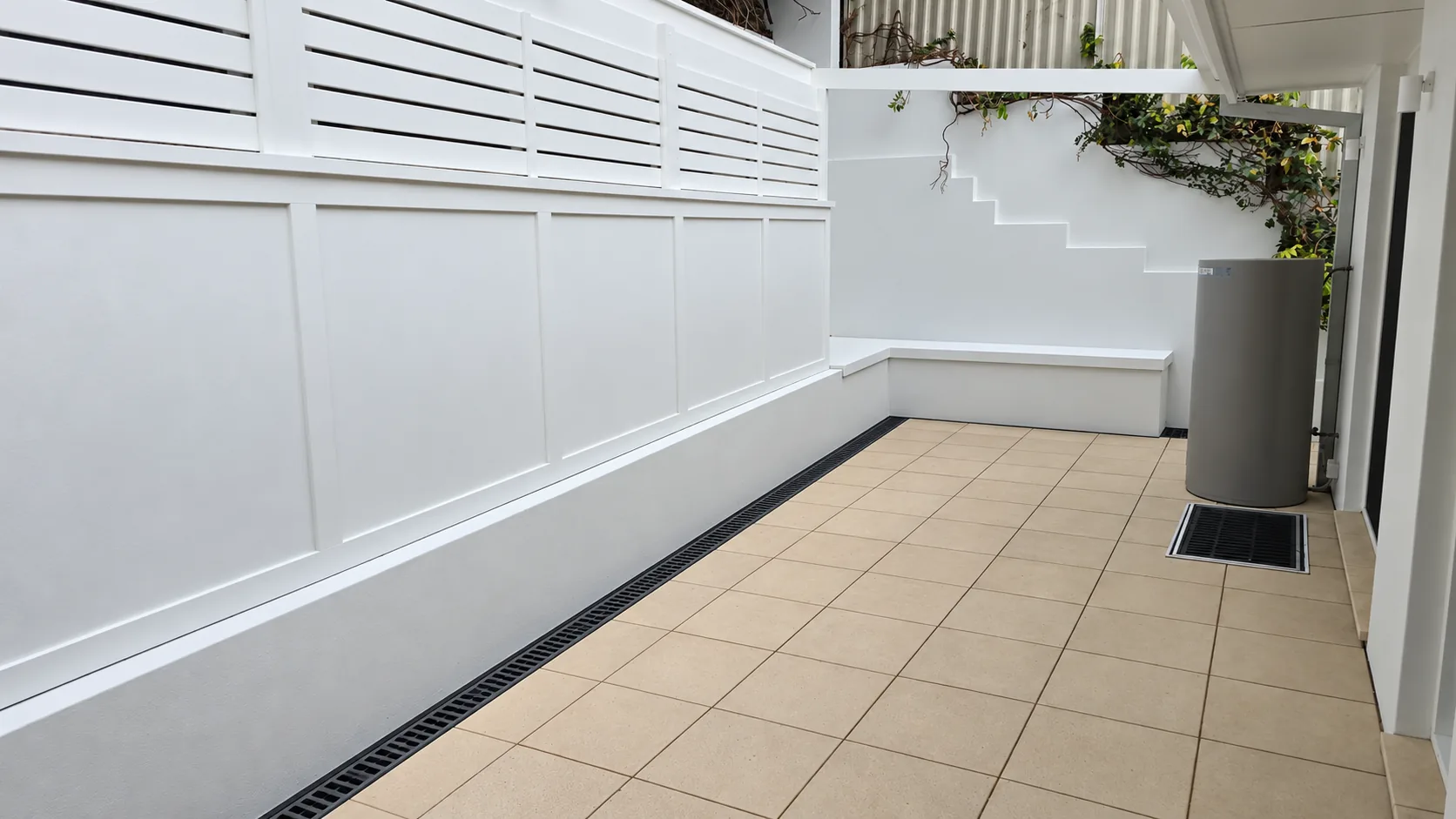 Private courtyard at the Terrigal residence, with crisp white panelled walls and slatted screening, sand-coloured paving edged by a linear drain, a built-in ledge and a water tank, with climbing greenery above