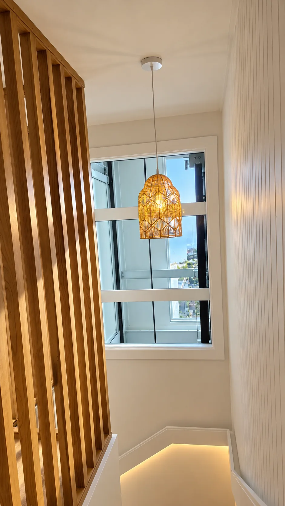 Stairwell with a woven pendant light hanging before a tall window, a screen of vertical timber battens and lit handrail detailing on the flight below