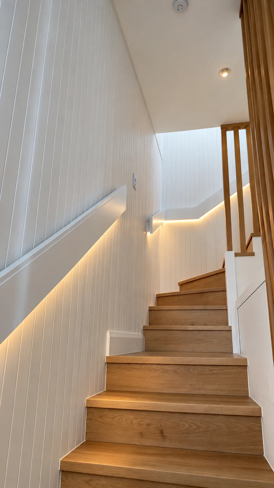 Timber staircase rising between vertically panelled white walls, with a warm strip of light concealed beneath the handrail and timber battens along the open side