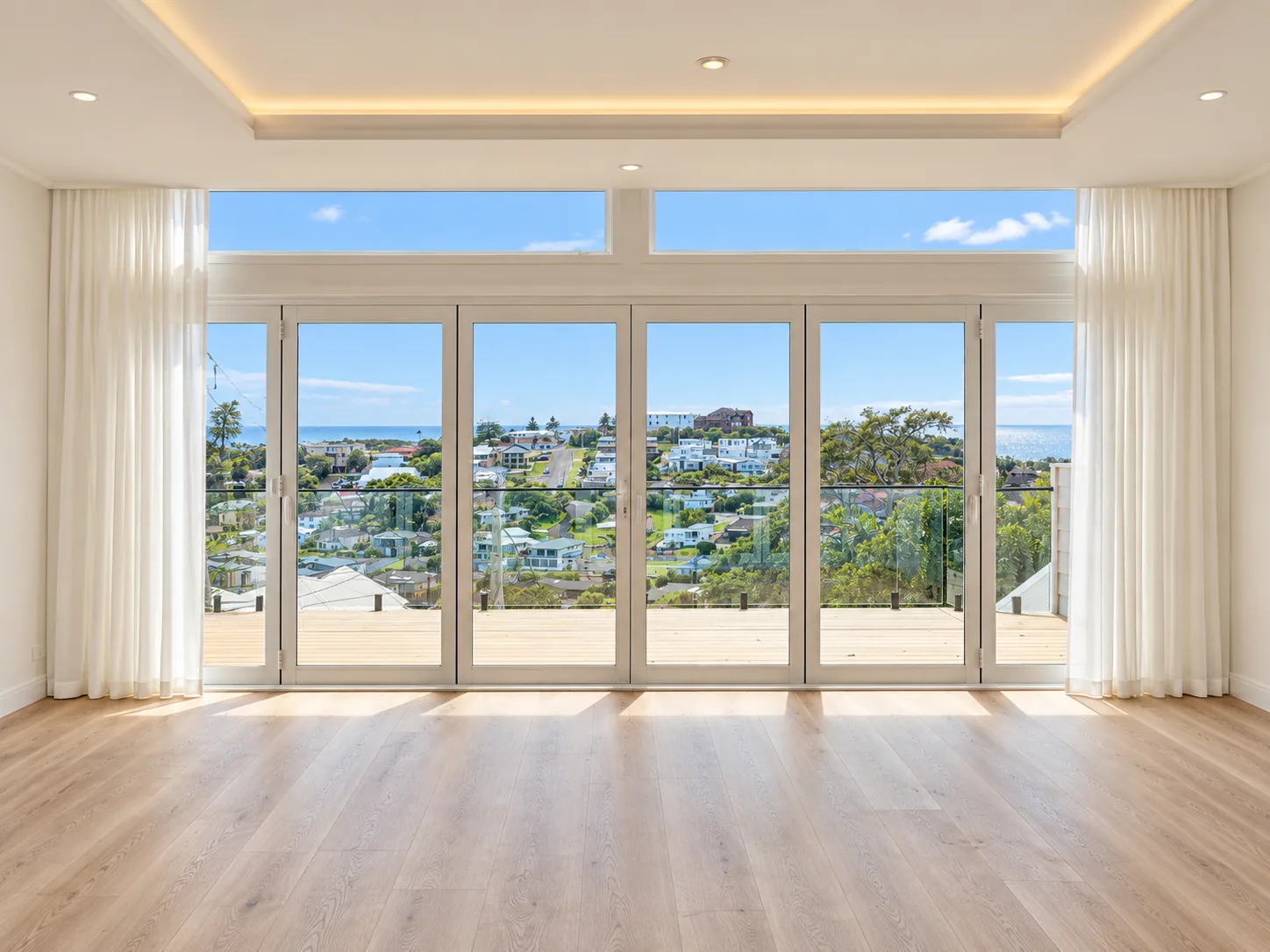 Sunlit Terrigal living room with a wall of glazed doors and highlight windows beneath a softly lit ceiling, sheer white curtains, and a timber deck with glass balustrade overlooking rooftops to the ocean